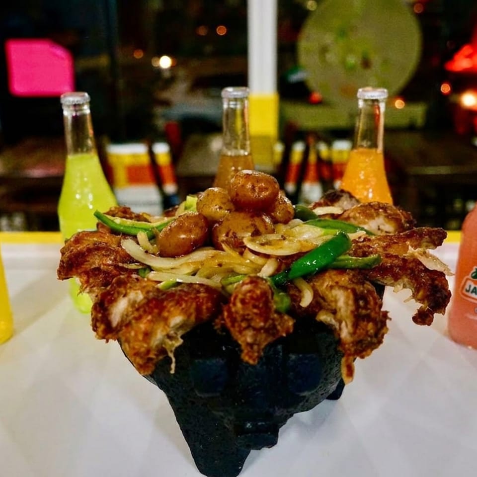Loaded platter of fried chicken, potatoes, peppers, and onions served with colorful Jarritos soda bottles in a restaurant.